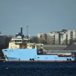 Γαλάζιο πλοίο υποστήριξης υπεράκτιων εργασιών με το όνομα Maersk Tracer δεμένο σε λιμάνι, με κοντέινερ και κτίρια στο βάθος