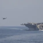 Το αεροπλανοφόρο USS Dwight D. Eisenhower πλέει σε θάλασσα με ομίχλη, με ελικόπτερο στον αέρα και πολεμικό πλοίο στο βάθος