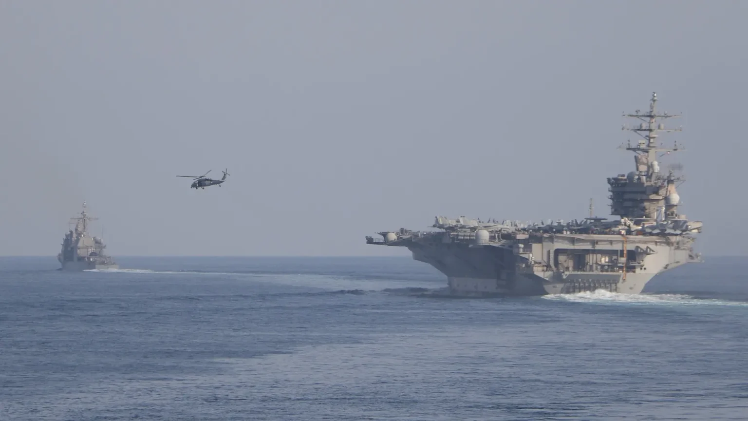 Το αεροπλανοφόρο USS Dwight D. Eisenhower πλέει σε θάλασσα με ομίχλη, με ελικόπτερο στον αέρα και πολεμικό πλοίο στο βάθος