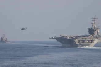 Το αεροπλανοφόρο USS Dwight D. Eisenhower πλέει σε θάλασσα με ομίχλη, με ελικόπτερο στον αέρα και πολεμικό πλοίο στο βάθος