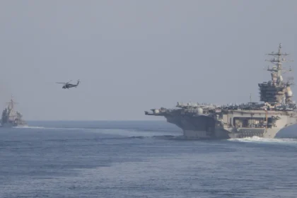 Το αεροπλανοφόρο USS Dwight D. Eisenhower πλέει σε θάλασσα με ομίχλη, με ελικόπτερο στον αέρα και πολεμικό πλοίο στο βάθος