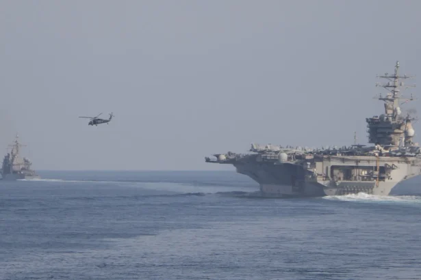 Το αεροπλανοφόρο USS Dwight D. Eisenhower πλέει σε θάλασσα με ομίχλη, με ελικόπτερο στον αέρα και πολεμικό πλοίο στο βάθος