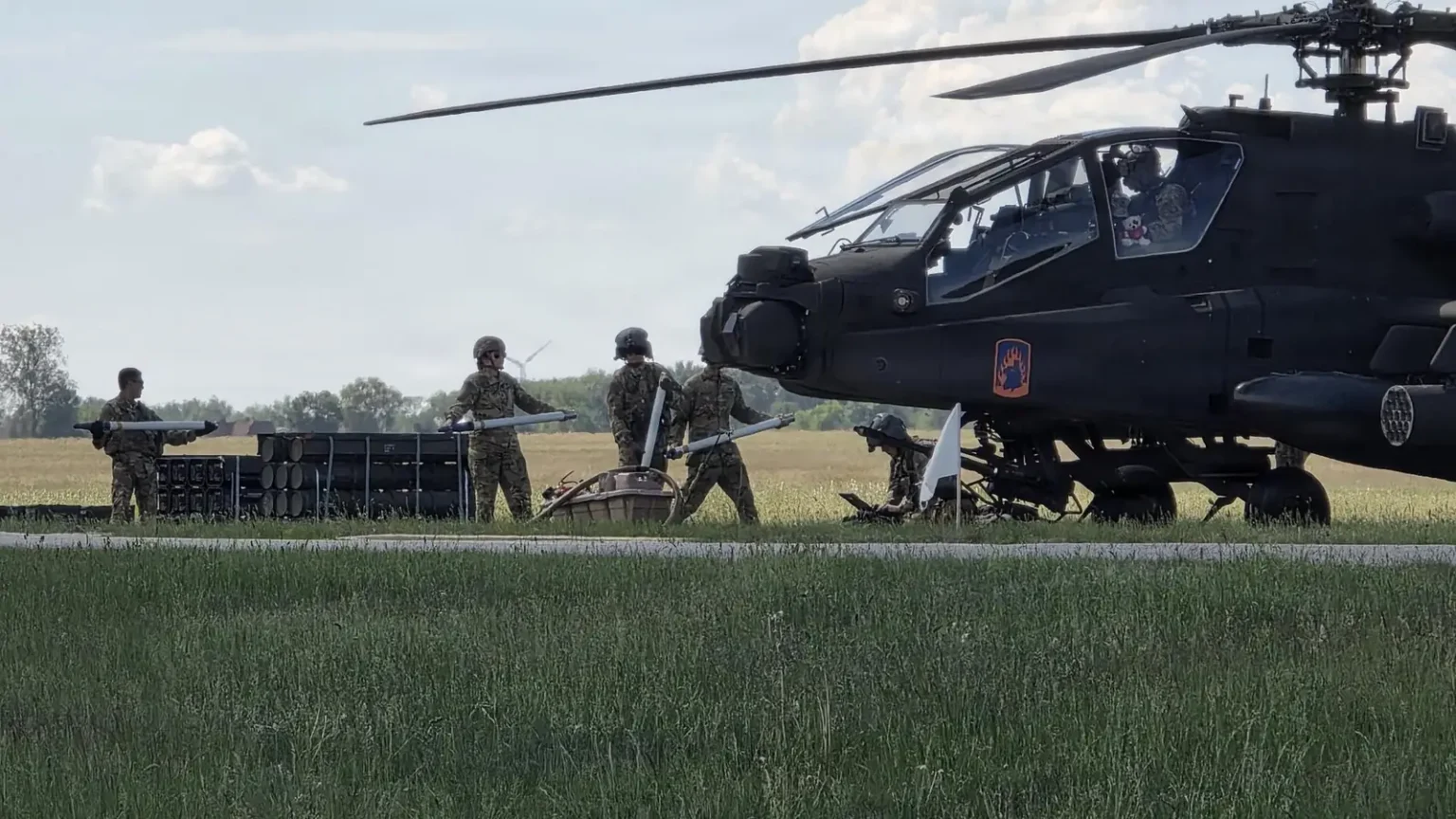 Επιθετικό ελικόπτερο AH-64E Apache σταθμευμένο σε χορτολιβαδική έκταση, με στρατιώτες να το προετοιμάζουν