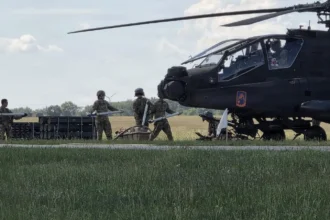 Επιθετικό ελικόπτερο AH-64E Apache σταθμευμένο σε χορτολιβαδική έκταση, με στρατιώτες να το προετοιμάζουν