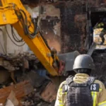 Διασώστης με κράνος μπροστά σε εκσκαφέα που απομακρύνει μπάζα από κατεστραμμένη πολυκατοικία