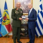 Ο Αρχηγός ΓΕΕΘΑ με στολή και ο γερουσιαστής Ρικ Σκοτ με μπλε κοστούμι κρατούν αναμνηστικό, μπροστά στις σημαίες του ΝΑΤΟ, της Ελλάδας και της ΕΕ