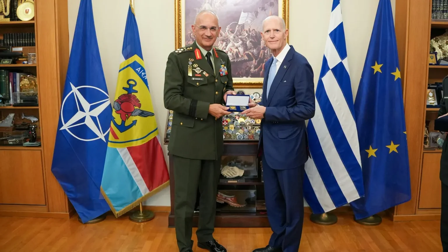 Ο Αρχηγός ΓΕΕΘΑ με στολή και ο γερουσιαστής Ρικ Σκοτ με μπλε κοστούμι κρατούν αναμνηστικό, μπροστά στις σημαίες του ΝΑΤΟ, της Ελλάδας και της ΕΕ