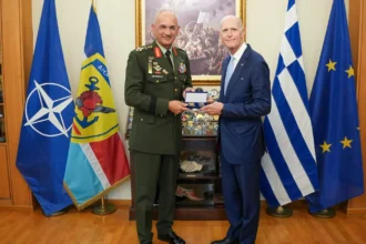 Ο Αρχηγός ΓΕΕΘΑ με στολή και ο γερουσιαστής Ρικ Σκοτ με μπλε κοστούμι κρατούν αναμνηστικό, μπροστά στις σημαίες του ΝΑΤΟ, της Ελλάδας και της ΕΕ