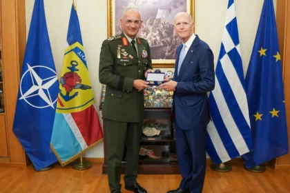 Ο Αρχηγός ΓΕΕΘΑ με στολή και ο γερουσιαστής Ρικ Σκοτ με μπλε κοστούμι κρατούν αναμνηστικό, μπροστά στις σημαίες του ΝΑΤΟ, της Ελλάδας και της ΕΕ