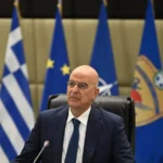 Ο υπουργός Εθνικής Άμυνας Νίκος Δένδιας καθισμένος μπροστά σε ελληνικές σημαίες και σημαίες ΕΕ και ΝΑΤΟ