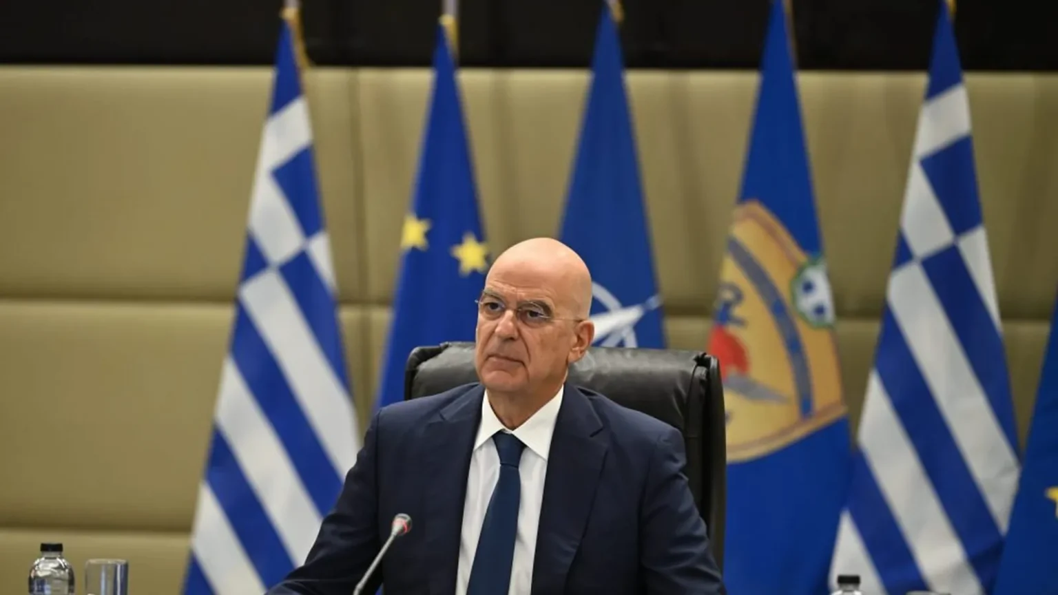 Ο υπουργός Εθνικής Άμυνας Νίκος Δένδιας καθισμένος μπροστά σε ελληνικές σημαίες και σημαίες ΕΕ και ΝΑΤΟ