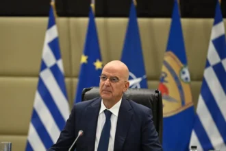 Ο υπουργός Εθνικής Άμυνας Νίκος Δένδιας καθισμένος μπροστά σε ελληνικές σημαίες και σημαίες ΕΕ και ΝΑΤΟ