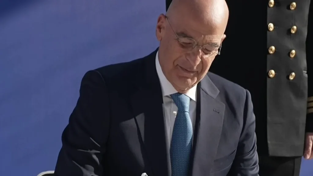 Ο υπουργός Εθνικής Άμυνας Νίκος Δένδιας υπογράφει καθιστός κατά την τελετή παράδοσης της φρεγάτας Νέαρχος, με αξιωματικό του Πολεμικού Ναυτικού πίσω του