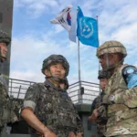 Αξιωματικοί της Νότιας Κορέας και των ΗΠΑ με κράνη συνομιλούν σε φυλάκιο, με τις σημαίες της Νότιας Κορέας και του ΟΗΕ στο βάθος
