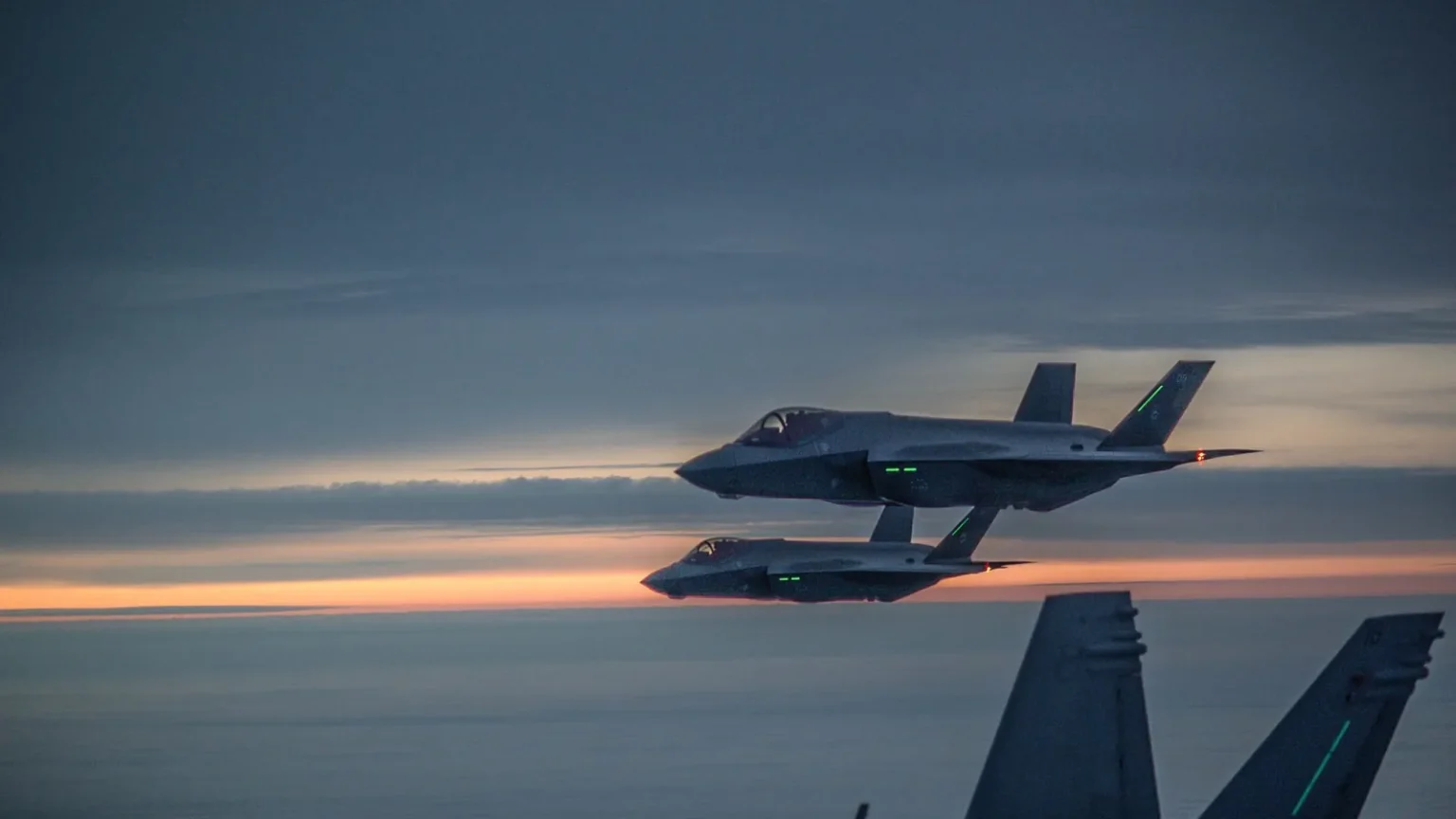 Δύο φινλανδικά F-35A πετούν σε σχηματισμό στο σούρουπο, με την ουρά ενός F/A-18 Hornet σε πρώτο πλάνο