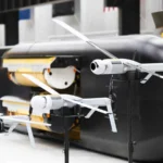 Λευκά drones ALTIUS με ανοιχτές έλικες σε βάσεις μπροστά σε μαύρο υποβρύχιο όχημα Ghost Shark με ανοιχτό χώρο φορτίου