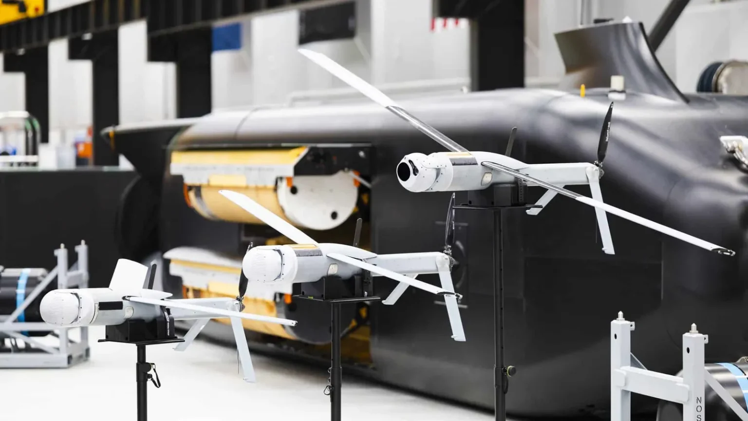 Λευκά drones ALTIUS με ανοιχτές έλικες σε βάσεις μπροστά σε μαύρο υποβρύχιο όχημα Ghost Shark με ανοιχτό χώρο φορτίου