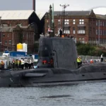 Το πυρηνοκίνητο υποβρύχιο HMS Agamemnon κλάσης Astute στο λιμάνι του Μπάροου-ιν-Φέρνες, με μέλη του πληρώματος στο κατάστρωμα και ρυμουλκό δίπλα του