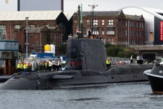 Το πυρηνοκίνητο υποβρύχιο HMS Agamemnon κλάσης Astute στο λιμάνι του Μπάροου-ιν-Φέρνες, με μέλη του πληρώματος στο κατάστρωμα και ρυμουλκό δίπλα του