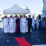 Ναυτικοί αξιωματικοί με λευκές στολές χαιρετούν στο κατάστρωμα πολεμικού πλοίου δίπλα στη σημαία της επιχείρησης IRINI