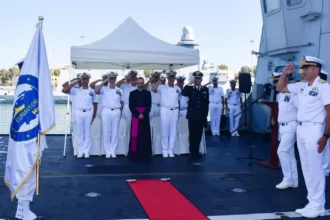 Ναυτικοί αξιωματικοί με λευκές στολές χαιρετούν στο κατάστρωμα πολεμικού πλοίου δίπλα στη σημαία της επιχείρησης IRINI