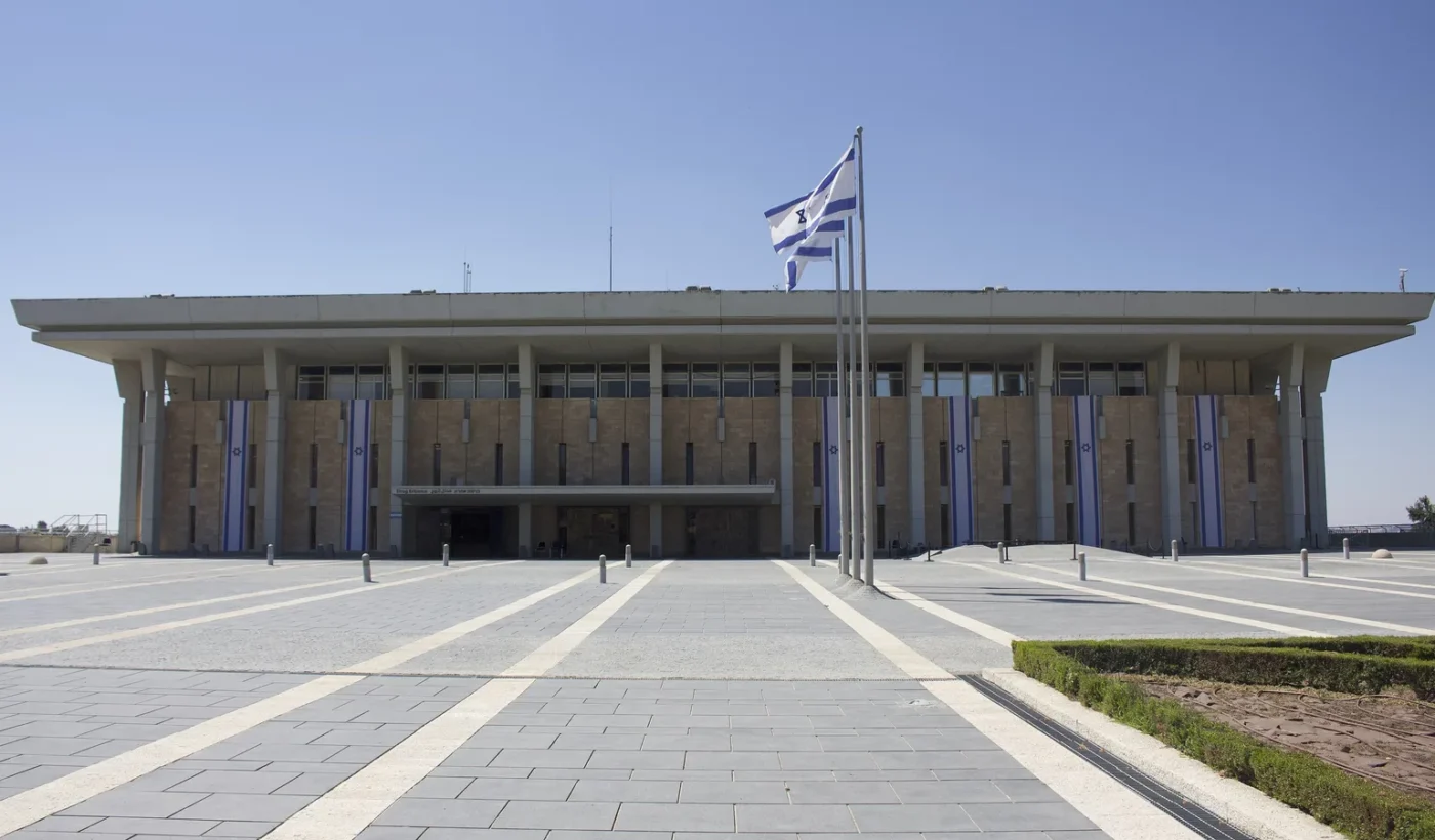 Η πρόσοψη του κτιρίου της Κνεσέτ στην Ιερουσαλήμ με την ισραηλινή σημαία σε ιστό και κάθετες σημαίες στις κολόνες, κάτω από καθαρό ουρανό