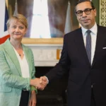 Ο Κωνσταντίνος Κόμπος με γυαλιά και σκούρο κοστούμι σφίγγει το χέρι της Ιβέτ Κούπερ σε αίθουσα με πράσινους τοίχους και βρετανική σημαία