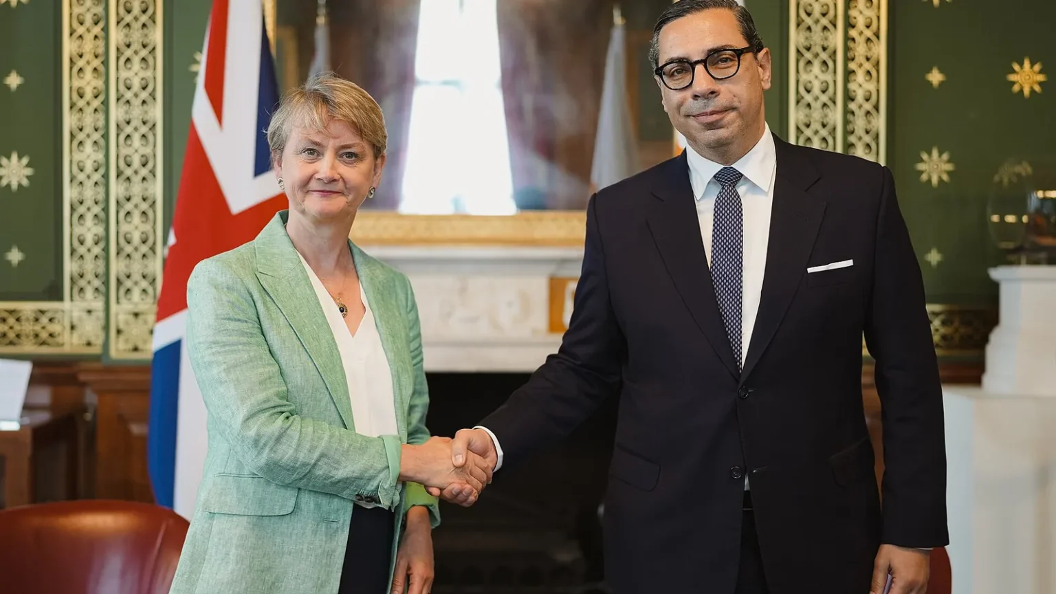 Ο Κωνσταντίνος Κόμπος με γυαλιά και σκούρο κοστούμι σφίγγει το χέρι της Ιβέτ Κούπερ σε αίθουσα με πράσινους τοίχους και βρετανική σημαία