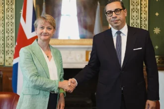 Ο Κωνσταντίνος Κόμπος με γυαλιά και σκούρο κοστούμι σφίγγει το χέρι της Ιβέτ Κούπερ σε αίθουσα με πράσινους τοίχους και βρετανική σημαία