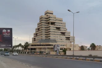 Ψηλό κλιμακωτό κτίριο σε φόντο συννεφιασμένου ουρανού δίπλα σε λεωφόρο με φοίνικες και φανοστάτες