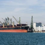 Φορτηγό πλοίο με κόκκινη γάστρα δεμένο κάτω από γερανούς στο λιμάνι της Βάρνας, με κτίρια της πόλης στο βάθος