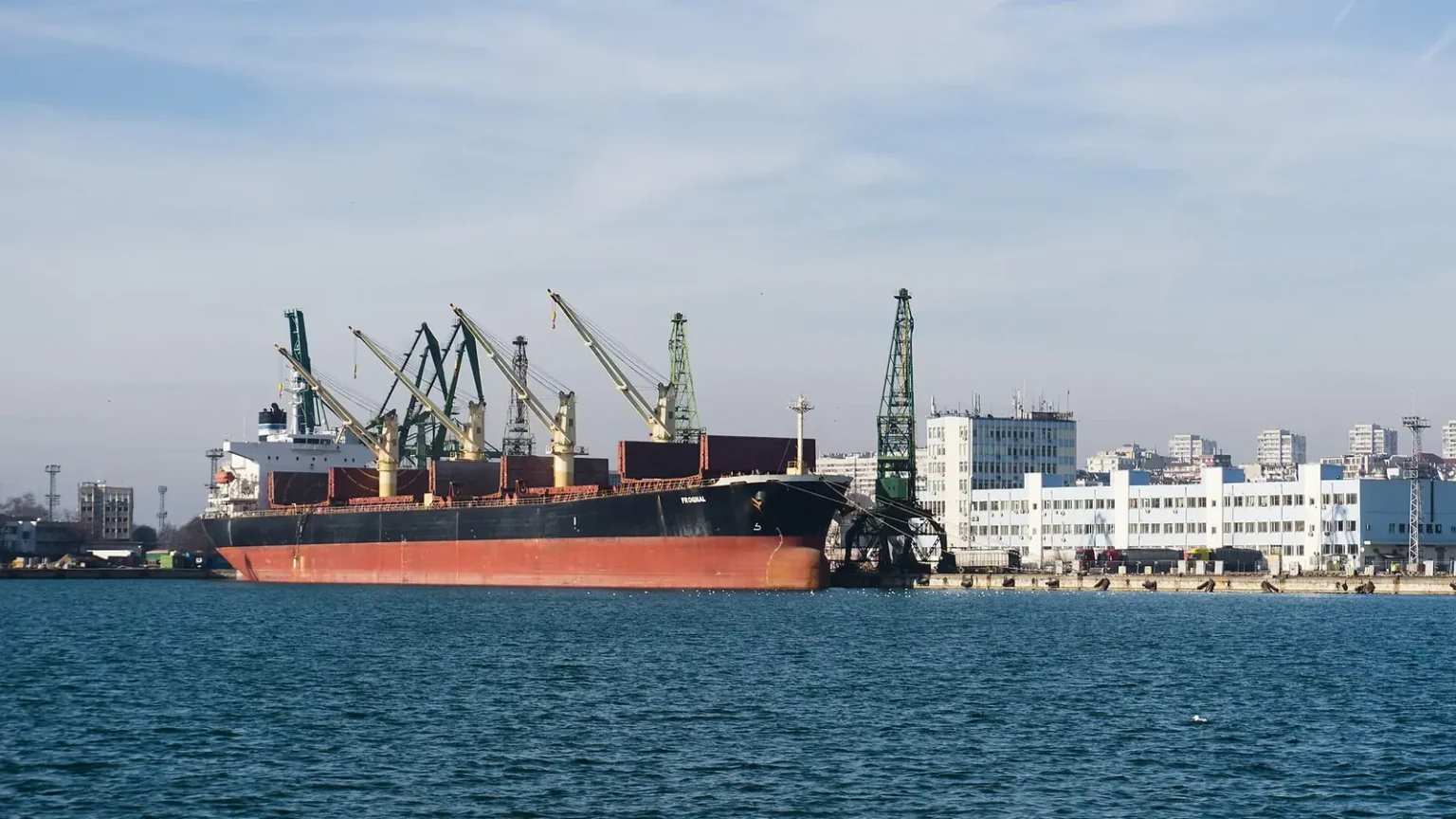 Φορτηγό πλοίο με κόκκινη γάστρα δεμένο κάτω από γερανούς στο λιμάνι της Βάρνας, με κτίρια της πόλης στο βάθος