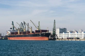Φορτηγό πλοίο με κόκκινη γάστρα δεμένο κάτω από γερανούς στο λιμάνι της Βάρνας, με κτίρια της πόλης στο βάθος