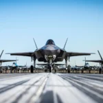 Τρία μαχητικά F-35A σε πρώτο πλάνο σε διάδρομο, με σειρές μαχητικών στο βάθος