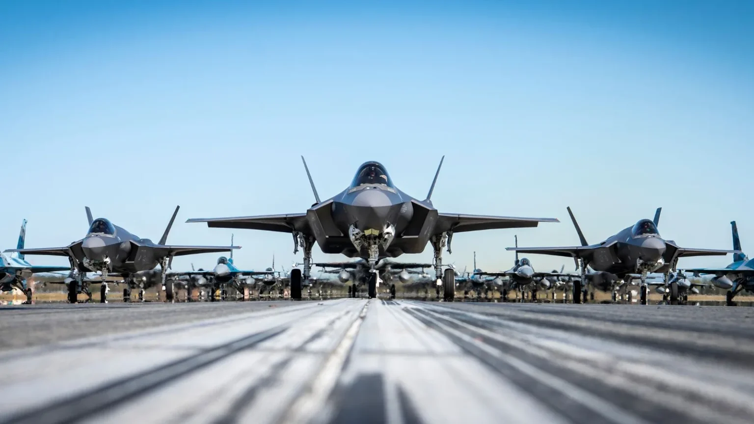 Τρία μαχητικά F-35A σε πρώτο πλάνο σε διάδρομο, με σειρές μαχητικών στο βάθος