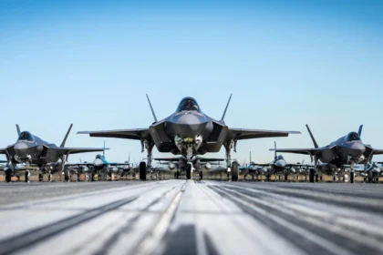 Τρία μαχητικά F-35A σε πρώτο πλάνο σε διάδρομο, με σειρές μαχητικών στο βάθος