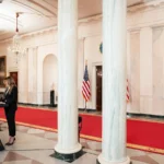 Η Μελάνια Τραμπ στο βήμα στον διάδρομο του Λευκού Οίκου με κόκκινο χαλί και μαρμάρινους κίονες