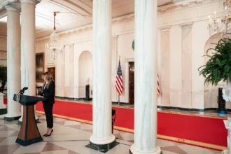Η Μελάνια Τραμπ στο βήμα στον διάδρομο του Λευκού Οίκου με κόκκινο χαλί και μαρμάρινους κίονες