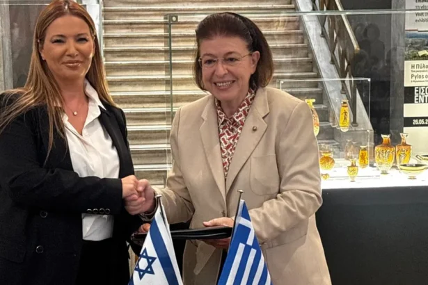 Η υπουργός Πολιτισμού Λίνα Μενδώνη και η πρέσβης του Ισραήλ Pninat Yanay σφίγγουν τα χέρια πίσω από τραπέζι με ισραηλινή και ελληνική σημαία στο Εθνικό Αρχαιολογικό Μουσείο