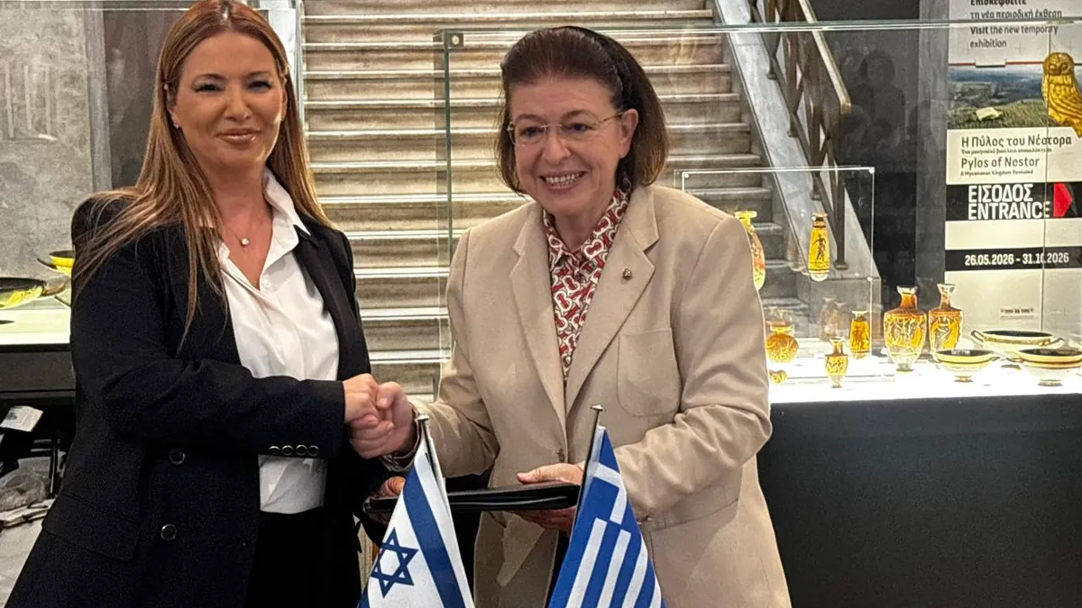 Η υπουργός Πολιτισμού Λίνα Μενδώνη και η πρέσβης του Ισραήλ Pninat Yanay σφίγγουν τα χέρια πίσω από τραπέζι με ισραηλινή και ελληνική σημαία στο Εθνικό Αρχαιολογικό Μουσείο