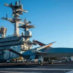 Γκρι μη επανδρωμένο αεροσκάφος MQ-25 στο κατάστρωμα πτήσεων αεροπλανοφόρου με ναύτες γύρω του