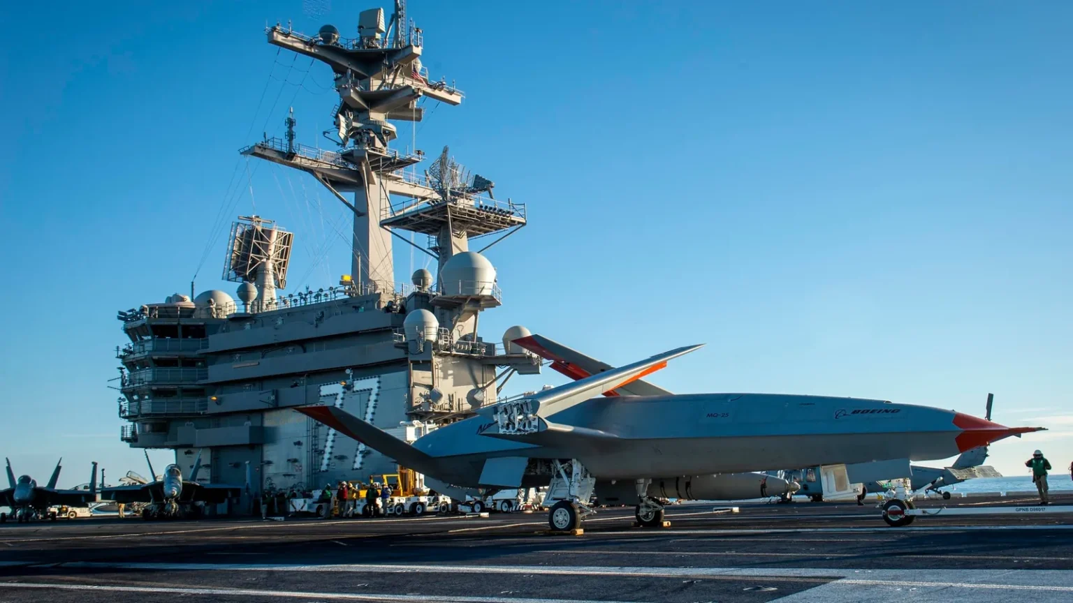 Γκρι μη επανδρωμένο αεροσκάφος MQ-25 στο κατάστρωμα πτήσεων αεροπλανοφόρου με ναύτες γύρω του