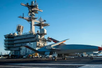 Γκρι μη επανδρωμένο αεροσκάφος MQ-25 στο κατάστρωμα πτήσεων αεροπλανοφόρου με ναύτες γύρω του