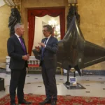 Δύο άνδρες με κοστούμια συζητούν σε αίθουσα με κόκκινο χαλί, δίπλα σε εκτιθέμενα μη επανδρωμένα αεροσκάφη