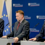 Ο υπουργός Εσωτερικών της Πολωνίας Μάρτσιν Κιερβίνσκι ανάμεσα στον υπουργό Τόμας Σιεμόνιακ και στον αρχηγό της Αστυνομίας Μάρεκ Μπορόν σε βήμα με σημαίες Πολωνίας και ΕΕ