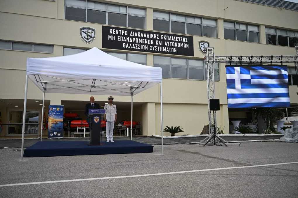 Ο υπουργός Εθνικής Άμυνας στο βήμα κάτω από λευκή τέντα, μπροστά στο κτίριο με την πινακίδα του νέου Κέντρου