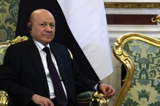 Ο Ρασάντ αλ Αλίμι καθισμένος μπροστά από τη σημαία της Υεμένης, με ακουστικό διερμηνείας