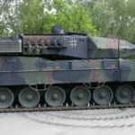 Πλάγια όψη άρματος μάχης Leopard 2A7 με παραλλαγή σε άσφαλτο, με δέντρα στο φόντο