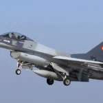 Γκρι μαχητικό F-16 με το ρουμανικό σήμα στο κάθετο σταθερό, σε πτήση με κατεβασμένα σκέλη προσγείωσης