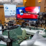 Τραπέζι με διάφορα drones πολλαπλών ελίκων και FPV, οθόνη με τη σημαία της Ταϊβάν και πανό Blue UAS
